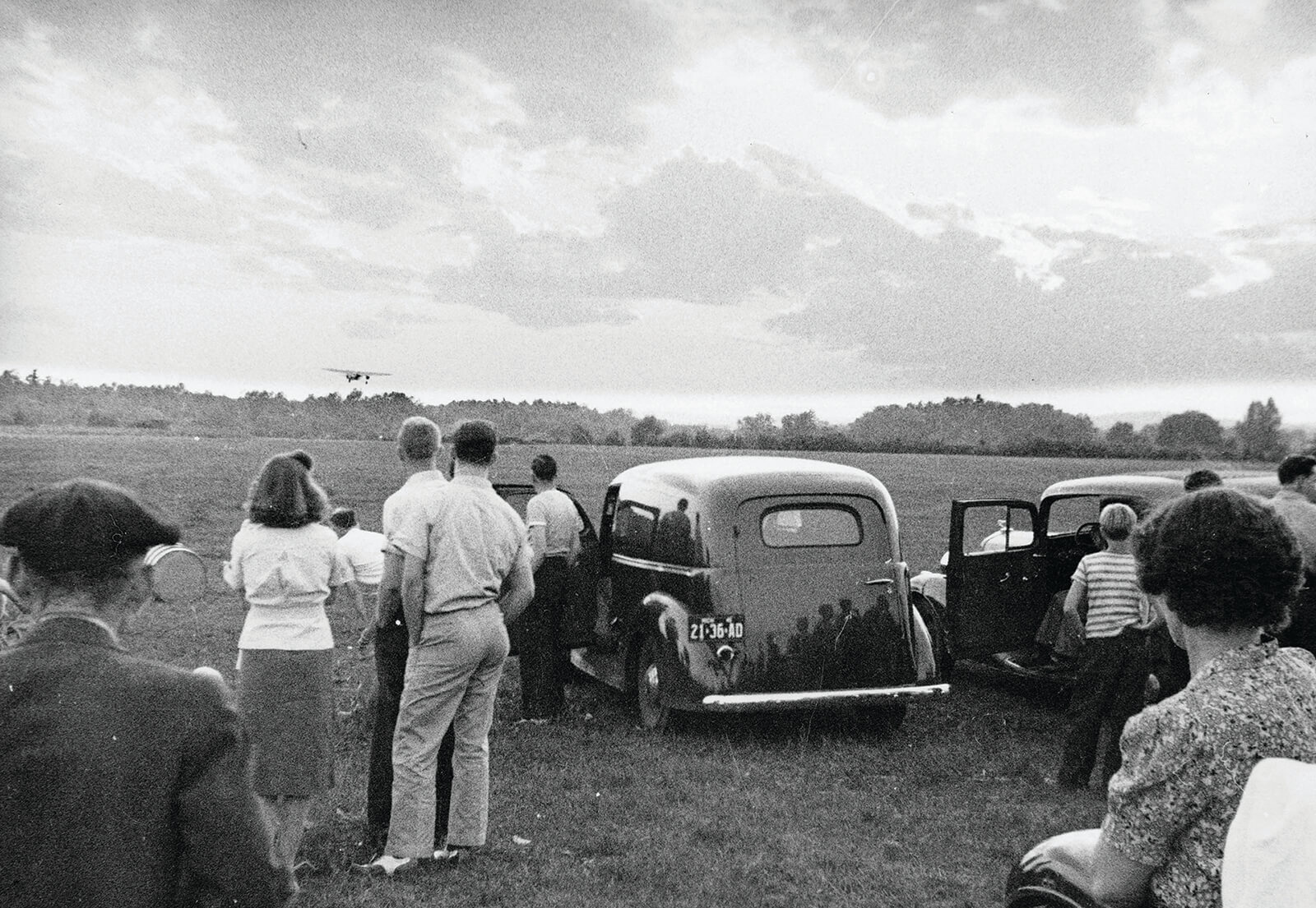 Past photo of people by old cars watching a plane fly low over an open field